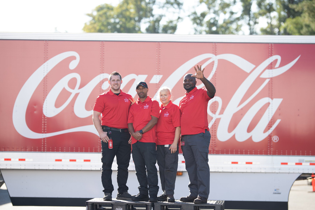 Four employees in red polo shirts in front of a large Coca-Cola sign
